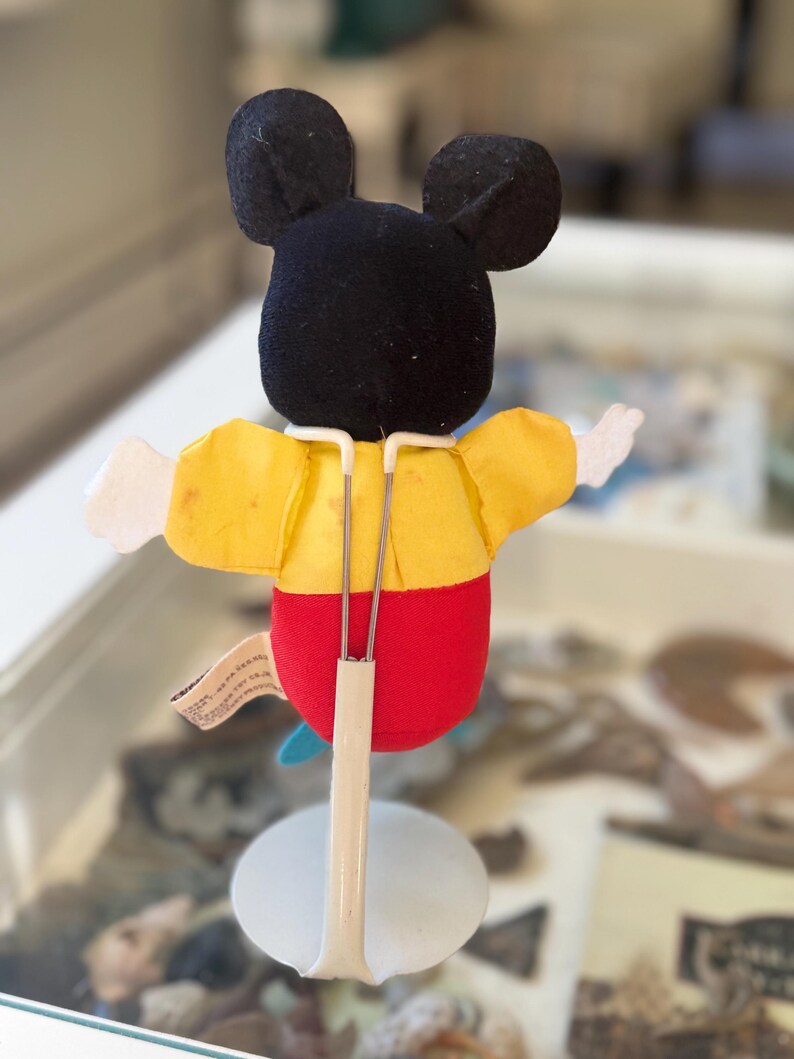 May include: A plush Mickey Mouse character figure viewed from the back, featuring a black head with round ears, a yellow shirt, and red pants. The figure is supported by a white metal display stand with two thin prongs holding the torso.