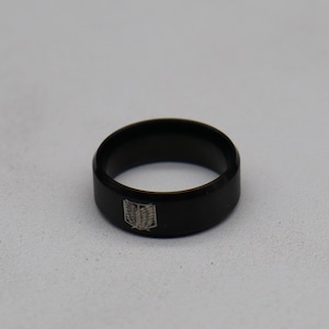 May include: A black ring with a flat band. The ring features a silver emblem on the front. The ring is made of a shiny, dark material. The ring is a piece of jewelry.