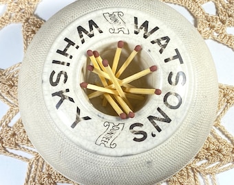 Puede incluir: Un cenicero redondo de cerámica blanquecina con las palabras "Whisky Watsons Dundee" impresas en negro. El cenicero contiene varias cerillas de madera con puntas rojas. El cenicero está sobre un tapete de ganchillo beige.