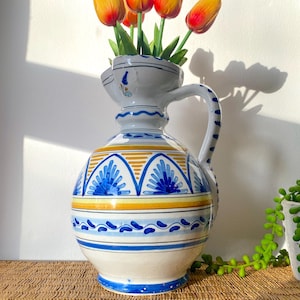 May include: A decorative ceramic pitcher with a bulbous body and a handle, adorned with blue and yellow geometric patterns. The pitcher holds several orange and yellow tulips. The pitcher is approximately 30 cm tall.