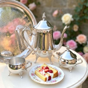 May include: A polished silver tea set, comprising a teapot, creamer, sugar bowl, and a serving tray, is displayed on a white table. A slice of cake with strawberries is on a floral plate. The backdrop is a garden with pink roses.