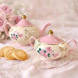 May include: A vintage-style tea set featuring a teapot and sugar bowl, decorated with pink roses and gold detailing. The teapot and sugar bowl have pink lids and handles. Several golden biscuits are arranged in front of the tea set.
