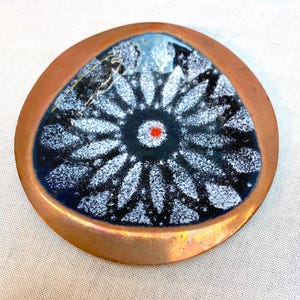 May include: A copper-coloured, triangular dish with a dark blue interior. The dish features a white and black floral design with a red centre. The rim of the dish is a warm copper colour.