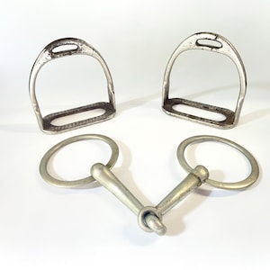 May include: A pair of silver-toned metal horse stirrups with textured footbeds and a matching silver-toned metal jointed snaffle bit with two circular rings, all featuring a weathered, metallic finish and a sturdy, functional construction.