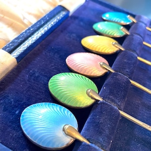 May include: Set of six silver teaspoons with enamel handles in shades of blue, green, yellow, and pink. The handles are decorated with a swirling pattern. The spoons are in a blue velvet box with the word "ENGLAND" engraved on one of the spoons.