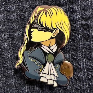 Może przedstawiać: Metalowa emaliowana przypinka przedstawiająca profil osoby z żółtymi włosami, opaską na oczy i niebieską kurtką z białymi falbanami. Przypinka ma złoty kontur i brązowy element na ramieniu. Tło to ciemnoszara dzianina.