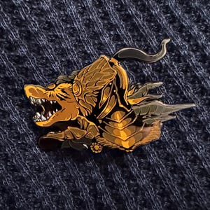Może przedstawiać: Szczegółowa emaliowana przypinka przedstawiająca stylizowane, złote zwierzę z ostrymi zębami i rogami. Przypinka ma skomplikowane detale i metaliczne wykończenie, na tle ciemnoniebieskiego, teksturowanego tła. Projekt zawiera elementy fantasy.