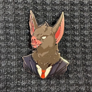 May include: A metal enamel pin featuring a stylised anthropomorphic bat with a pig-like snout, pointed ears with pink inner details, and white eyes. The creature is dressed in a dark navy blue suit jacket, a white collared shirt, and a red tie.