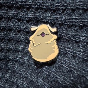 May include: A tan and cream-colored enamel pin in the shape of a fluffy, cartoonish creature with a purple mouth. The pin has a gold outline and is set against a dark blue knitted background.