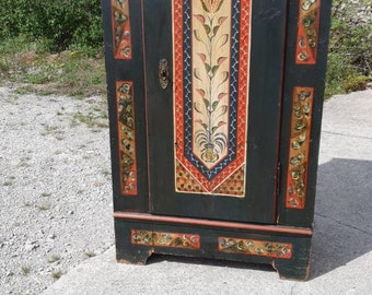 Pode incluir: Um armário de madeira pintado de preto com uma única porta. A porta é decorada com um desenho floral colorido em vermelho, amarelo, azul e verde. O armário tem um estilo simples e tradicional.