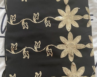 May include: Black fabric with gold sequin floral and vine patterns. The design features large, detailed flowers and delicate vines, creating an elegant and ornate look. The fabric is folded, showcasing the intricate sequin work.