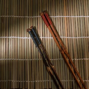 May include: Four pairs of chopsticks rest on a bamboo mat. Two pairs have dark brown handles wrapped with black thread. The other two pairs have light brown handles with red thread wrapping. The chopsticks are angled across the mat.