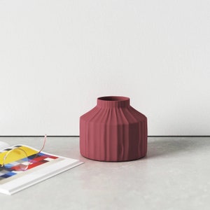 May include: A deep red ceramic vase with a ribbed design. The vase is sitting on a grey surface.