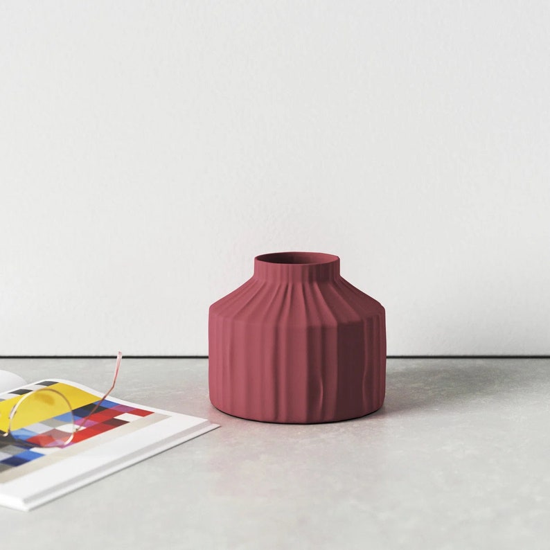 May include: A deep red ceramic vase with a ribbed design. The vase is sitting on a grey surface.