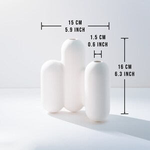 Może przedstawiać: Biały ceramiczny wazon o unikalnym, organicznym kształcie. Wazon ma 15 cm szerokości i 16 cm wysokości.