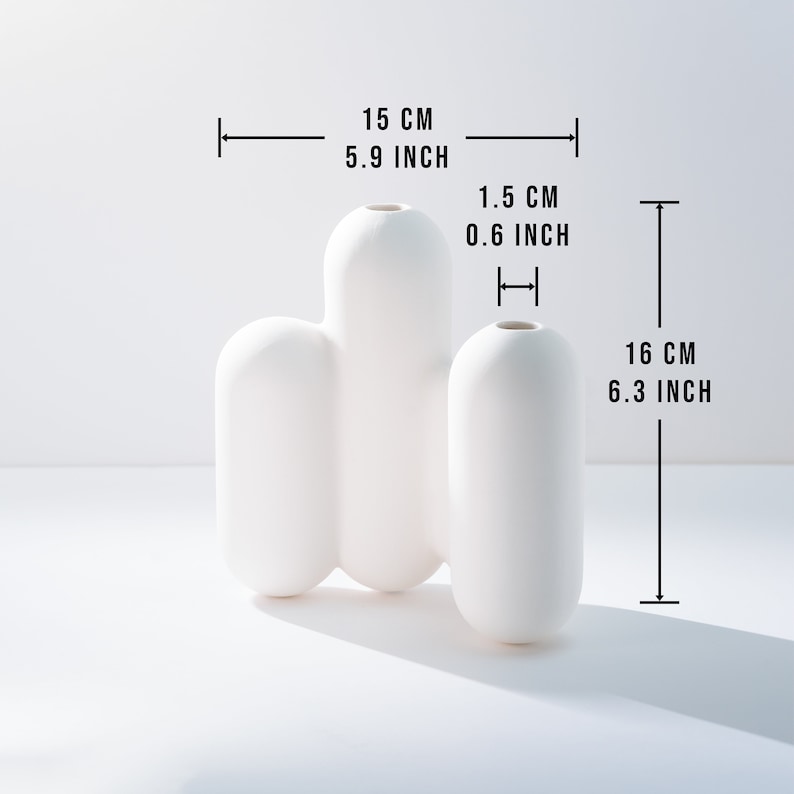 Może przedstawiać: Biały ceramiczny wazon o unikalnym, organicznym kształcie. Wazon ma 15 cm szerokości i 16 cm wysokości.