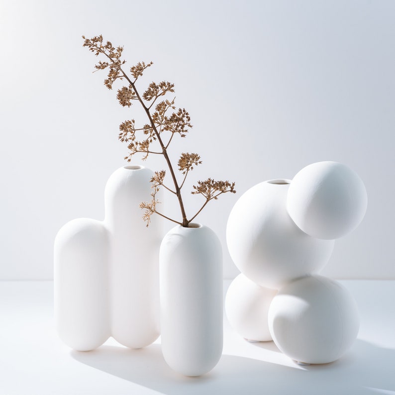 Może przedstawiać: Zestaw trzech białych ceramicznych wazon&oacute;w o organicznych, zaokrąglonych kształtach. Wazy są ułożone w grupie, z pojedynczą suchą gałązką kwiat&oacute;w w centralnym wazonie.