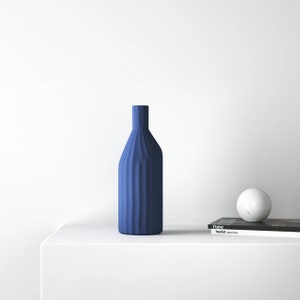 May include: A blue ceramic vase with a textured, ribbed design sits on a white surface. A white marble sphere and a magazine titled "Tourist Japan House" are also on the surface.