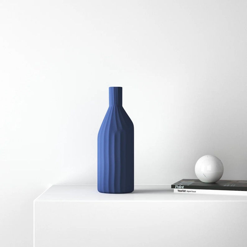 May include: A blue ceramic vase with a textured, ribbed design sits on a white surface. A white marble sphere and a magazine titled "Tourist Japan House" are also on the surface.