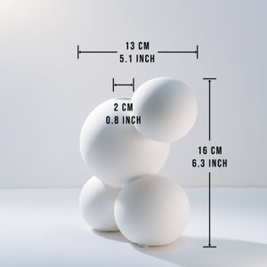 Może przedstawiać: Biały ceramiczny wazon o unikalnym, organicznym kształcie. Wazon składa się z czterech połączonych ze sobą kul, z otworem o średnicy 2 cm na g&oacute;rze. Wazon ma 13 cm szerokości i 16 cm wysokości.