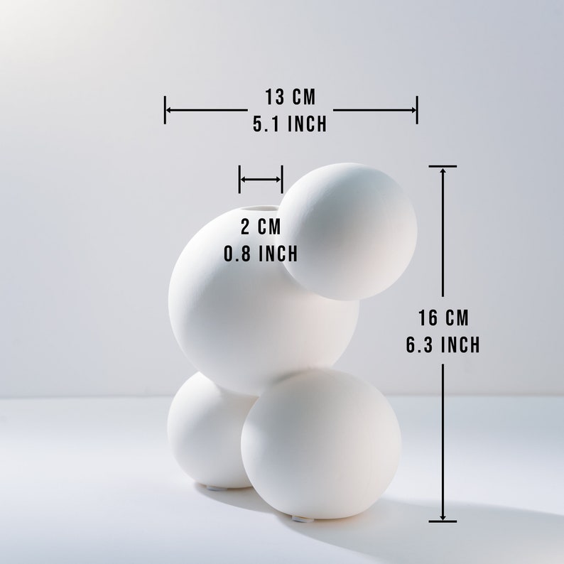 Może przedstawiać: Biały ceramiczny wazon o unikalnym, organicznym kształcie. Wazon składa się z czterech połączonych ze sobą kul, z otworem o średnicy 2 cm na g&oacute;rze. Wazon ma 13 cm szerokości i 16 cm wysokości.