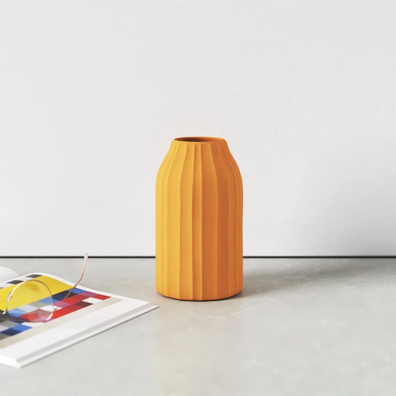 May include: A tall, yellow ceramic vase with a ribbed texture. The vase is empty and sitting on a light grey surface.