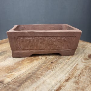 May include: A rectangular, reddish-brown bonsai pot with a textured surface and decorative carved design. The pot has a low profile with small feet and is made of a terracotta-like material. The pot is sitting on a wooden surface.