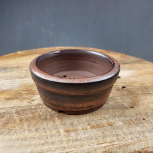 Puede incluir: Pequeño tiesto redondo de cerámica marrón para bonsái, con un borde ligeramente ensanchado. El tiesto tiene un aspecto texturizado y rústico con variaciones de color, desde marrón oscuro hasta marrón rojizo. Está sobre una superficie de madera.