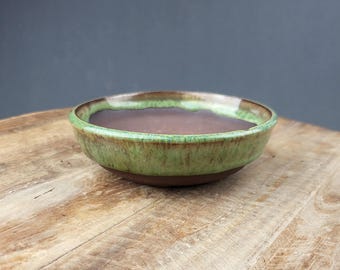 Puede incluir: Una maceta redonda de cerámica para bonsái con interior marrón y exterior verde y marrón. La maceta tiene un diseño ancho y poco profundo y se asienta sobre una superficie de madera.