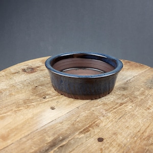 May include: A small, round ceramic bowl with a dark blue exterior and a brown interior. The bowl has a textured, ribbed design on the outside and sits on a wooden surface. The rim of the bowl is a lighter shade of blue.