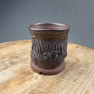 May include: A handcrafted ceramic cup with a textured, bark-like exterior. The cup features a dark brown rim and a base, with a lighter brown interior. The rough texture and earthy tones suggest a rustic, artisanal aesthetic, suitable for beverages or decorative use.