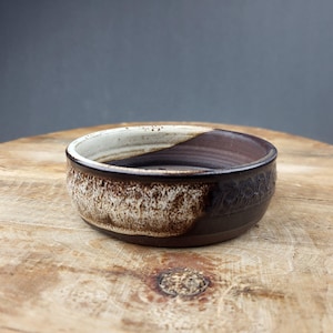 Könnte beinhalten: Eine handgefertigte Keramikschale mit zweifarbigem Design. Die obere Hälfte ist weiß und die untere Hälfte ist braun. Die Schale hat eine strukturierte Außenseite und eine glatte Innenseite. Die Schale steht auf einer Holzoberfläche.