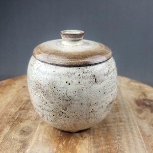 Puede incluir: Un tarro de cerámica redondo con tapa. El tarro es de color blanquecino con motas marrones y un borde marrón en la tapa. La tapa tiene un pequeño pomo. El tarro está sobre una superficie de madera.