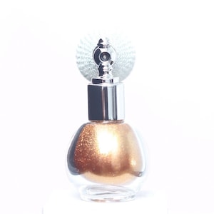 May include: A clear, round glass bottle with a silver-colored top and a decorative silver spray nozzle. The bottle is filled with a shimmering, golden-brown liquid. The background is white.