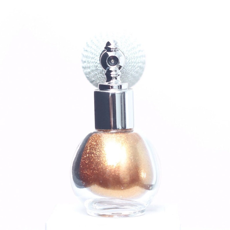 May include: A clear, round glass bottle with a silver-colored top and a decorative silver spray nozzle. The bottle is filled with a shimmering, golden-brown liquid. The background is white.