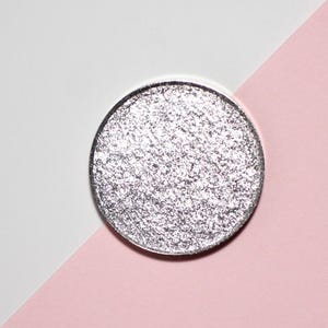 May include: A silver eyeshadow compact with a glittery, textured surface. The round compact is set against a backdrop of white and pink paper, creating a contrasting visual. The product is designed for eye makeup.