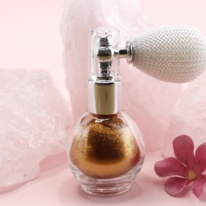 May include: A clear glass perfume bottle filled with gold glitter, topped with a silver and clear atomizer. The atomizer has a white, textured bulb. A pink flower and pink quartz rocks are in the background.