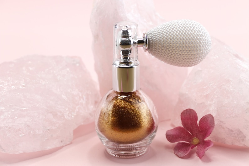 May include: A clear glass perfume bottle filled with gold glitter, topped with a silver and clear atomizer. The atomizer has a white, textured bulb. A pink flower and pink quartz rocks are in the background.