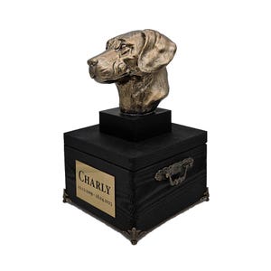 Op de afbeelding: Een zwarte houten urn voor huisdieren met een gouden metalen handvat en een bronzen hondenkopbeeld bovenop. De urn heeft een gouden plaquette met de naam "CHARLY" en de data "12.13.2009 - 18.04.2023" erop gegraveerd.