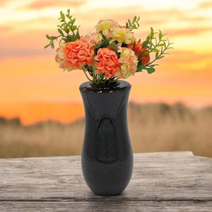 Könnte beinhalten: Eine schwarze Vase mit strukturierter Oberfläche hält einen Strauß aus orangefarbenen, gelben und rosa Blumen mit grünen Blättern. Die Vase steht auf einem Holztisch, mit einem verschwommenen Sonnenuntergang im Hintergrund.