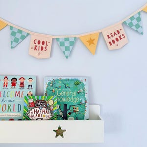 Puede incluir: Una guirnalda de tela con banderines que alternan entre motivos de estrellas amarillas y cuadros de color verde azulado. Dos banderines color crema muestran el texto rojo "COOL KIDS" y "READ BOOKS" con estrellas, colgados de un cordel claro.