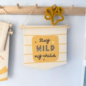 Può includere: Striscione color crema con la scritta "Stay WILD my child" in nero e giallo. Lo striscione è appeso a un'asta di legno con una decorazione a forma di impronta di zampa gialla sopra. Sono visibili anche altri striscioni con disegni di sole e luna.