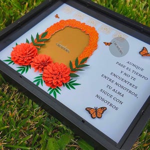 May include: A black shadow box frame containing a white background with three dimensional orange paper marigold flowers, green paper leaves, and monarch butterfly cutouts. The text reads: AUNQUE PASE EL TIEMPO Y NO TE ENCUENTRES ENTRE NOSOTROS, TU ALMA SIGUE CON NOSOTROS.