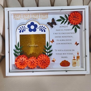 May include: A white shadow box frame containing a gold-colored photo frame, orange marigold paper flowers, butterflies, and candles. The text reads: AUNQUE PASE EL TIEMPO Y NO TE ENCUENTRES ENTRE NOSOTROS, TU ALMA SIGUE CON NOSOTROS. TE VOY A ELEGIR EN TODAS MIS VIDAS, MAMÁ.