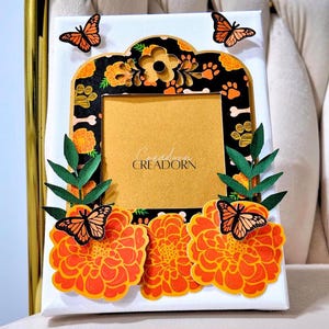 May include: A rectangular white canvas frame featuring a black border decorated with orange marigold flowers, paw prints, and bone shapes. The frame is adorned with green paper leaves and four monarch butterflies. The center displays the text CREADORN.