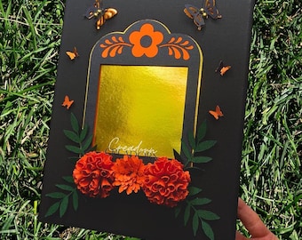May include: A rectangular black canvas featuring a central gold foil square, an orange floral arch, and orange butterfly cutouts. Three orange marigold-style flowers with dark green paper leaves are attached to the bottom of the frame.