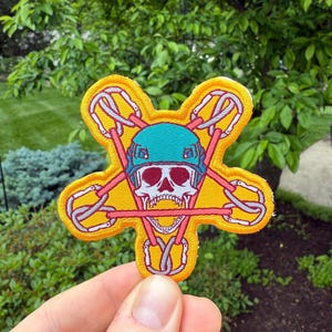 May include: Embroidered patch with a yellow starburst design. The patch features a skull wearing a teal helmet, surrounded by climbing carabiners. The skull has red eyes and a visible jaw. The patch is held in a hand against a background of green foliage.