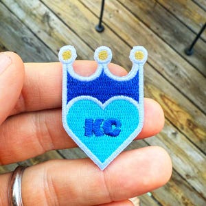 May include: Embroidered patch in the shape of a crown with a heart. The patch is blue and light blue with gold accents. The letters "KC" are embroidered on the heart.