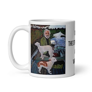 May include: White ceramic mug featuring a painting of a man in a boat with two dogs. The artwork depicts a man with a white beard and two dogs, one white and one brown and white. Text on the mug reads "The Other Side of the River".