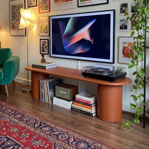 Può includere: Un soggiorno con una parete di galleria d'arte incorniciata e foto sopra una grande TV. Una console multimediale in stile moderno di metà secolo ospita un giradischi, libri e un altoparlante. Una poltrona in velluto verde e un tappeto fantasia completano lo spazio.